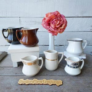 May include: A collection of seven ceramic creamers in various colors and patterns, including solid black, brown, and white, alongside a white vase holding a large pink flower. A wooden sign with the text Hudson River Craftings is in the foreground.