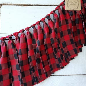 May include: A red and black buffalo plaid fabric garland with knotted ties. The frayed edges of the fabric create a rustic look. A small tag with the text "Hudson River Craftings HOME DECOR" is attached.