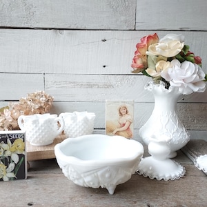 May include: White milk glass bowls, cups, and a vase with a floral design. The vase is filled with silk flowers in shades of pink and white. The items are arranged on a wooden surface with a vintage seed packet.