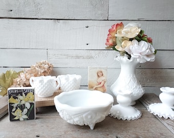 May include: White milk glass bowls, cups, and a vase with a floral design. The vase is filled with silk flowers in shades of pink and white. The items are arranged on a wooden surface with a vintage seed packet.