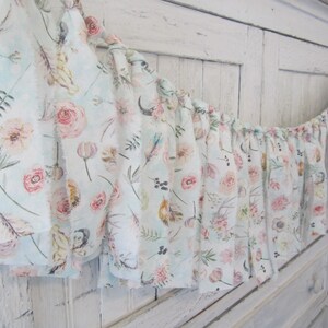 May include: A floral fabric garland with a ruffled edge. The fabric features a light blue background with pink roses, feathers, and other botanical elements. The garland is displayed against a white, distressed wooden surface.