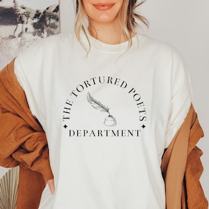May include: White sweatshirt with a black graphic design that says "The Tortured Poets Department" in a circle with a quill pen and inkwell in the center.