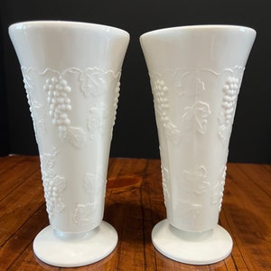 May include: Two white milk glass vases with a grapevine design. The vases have a footed base.