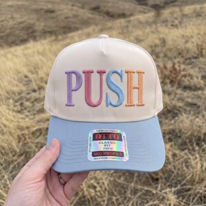 Op de afbeelding: Een crèmekleurige baseballpet met het woord PUSH geborduurd in paarse, roze, blauwe en oranje letters. De klep is lichtblauw en bevat een holografische sticker met de tekst OTTO, PREMIUM FABRIC, CLASSIC FIT, OSFM.