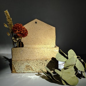 Könnte beinhalten: Ein beigefarbener Keramik-Wandtopf mit einem hausförmigen Oberteil und einem Loch zum Aufhängen. Der Topf hat eine gesprenkelte Textur und ist mit getrockneten Blumen und Grün gefüllt.