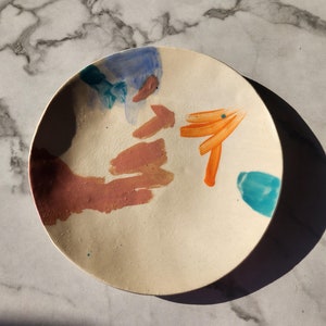Peut inclure: Une assiette en céramique blanche avec des coups de pinceau abstraits dans des tons d'orange, rose, bleu et turquoise.