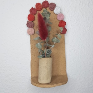 May include: A small, terracotta-colored wall planter with a rounded top and a shelf. The planter is decorated with a row of red, pink, and white flowers. A small vase with a dried red flower and green leaves sits on the shelf.