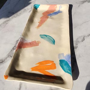 Pode incluir: Bandeja retangular de cerâmica creme com pinceladas abstratas em laranja, azul e rosa. A bandeja tem uma borda marrom escura e uma alça embutida. O design é moderno e artístico, ideal para servir.
