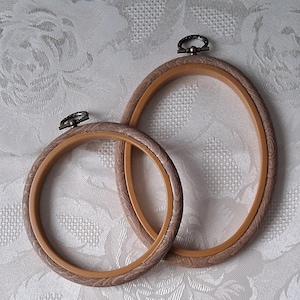 May include: Two oval embroidery hoops. The outer ring has a wood grain pattern, and the inner ring is a lighter tan colour. Each hoop has a metal clasp at the top for tightening the fabric. The hoops are overlapping on a white patterned surface.