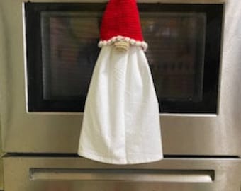 Peut inclure: Un porte-serviette en forme de gnome crocheté en rouge et blanc avec une barbe blanche et un nez brun. Le gnome est accroché à une porte de four en argent.