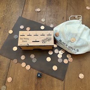 Pode incluir: Um jogo de caixa de madeira chamado "The Penny Game" com uma bandeja de dados e cinco ranhuras para centavos. A caixa é feita nos EUA e tem uma bolsa de cordão branco com o texto "BLACK HILLS GAME WORKS CO" impresso nela.