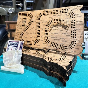 May include: A wooden game board shaped like the state of South Dakota. The board is made of multiple layers of wood and has a dark brown stain. The board has a grid of holes and the names of cities and towns in South Dakota are printed on the board. The board is stacked on top of other boards, which are also shaped like South Dakota.