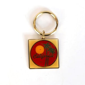 May include: A gold-colored keychain with a square pendant featuring a red circle with a yellow sun and green palm trees. The word "California" is written in cursive. The keychain has a gold ring.