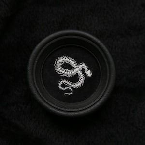 Può includere: Toppa circolare nera con un motivo a serpente ricamato in bianco. La toppa è posta su uno sfondo nero strutturato. Il serpente è dettagliato, con un corpo arrotolato e una testa curva.