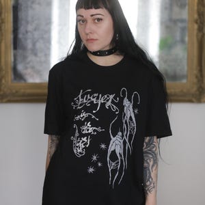 Op de afbeelding: Zwart T-shirt met een wit grafisch ontwerp met gestileerde tekst en abstracte vormen. Het shirt heeft korte mouwen en een ronde hals. Het model draagt een zwarte choker.