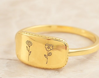 May include: A gold-colored ring with a rectangular face featuring two delicate flower illustrations. The ring's band is smooth, and the face has a beaded border. The flowers are etched in black, creating a contrast against the gold. Jewelry.