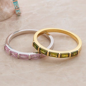 以下が含まれることがあります： カラフルな宝石がついた、重ね付けできる3つのリング。一番上のリングは緑色の宝石がついたゴールド、真ん中のリングはピンク色の宝石がついたシルバー、一番下のリングは青色の宝石がついたローズゴールドです。