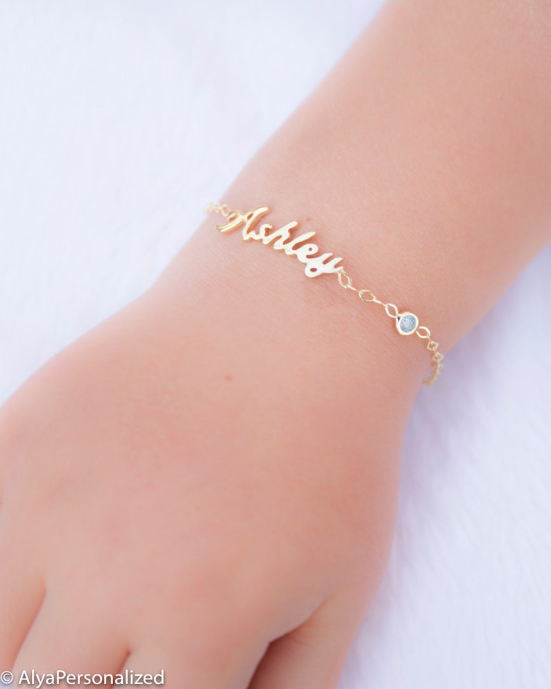 May include: Gold name bracelet with a blue gemstone charm. The name "Ashley" is written in cursive on the bracelet.