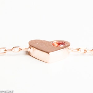 May include: A rose gold heart charm with a small red gemstone set in the center, attached to a delicate rose gold chain.