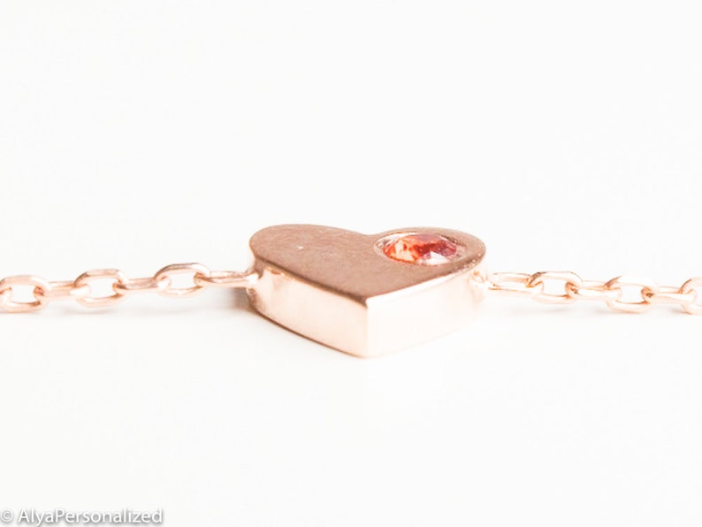May include: A rose gold heart charm with a small red gemstone set in the center, attached to a delicate rose gold chain.
