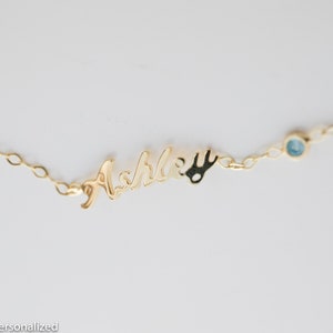 May include: Gold necklace with a cursive nameplate that reads "Ashley" and a small blue gemstone charm.