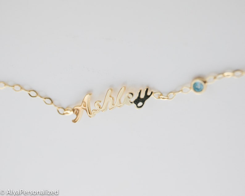 May include: Gold necklace with a cursive nameplate that reads "Ashley" and a small blue gemstone charm.