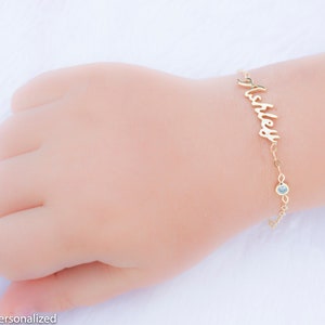 May include: Gold name bracelet with a single clear gemstone charm. The bracelet is personalized with the name "Ashley" in a cursive font.