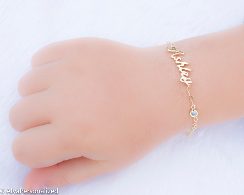 May include: Gold name bracelet with a single clear gemstone charm. The bracelet is personalized with the name "Ashley" in a cursive font.