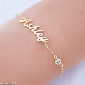 May include: Gold name bracelet with a light blue gemstone charm. The bracelet says "Ashley" in cursive.