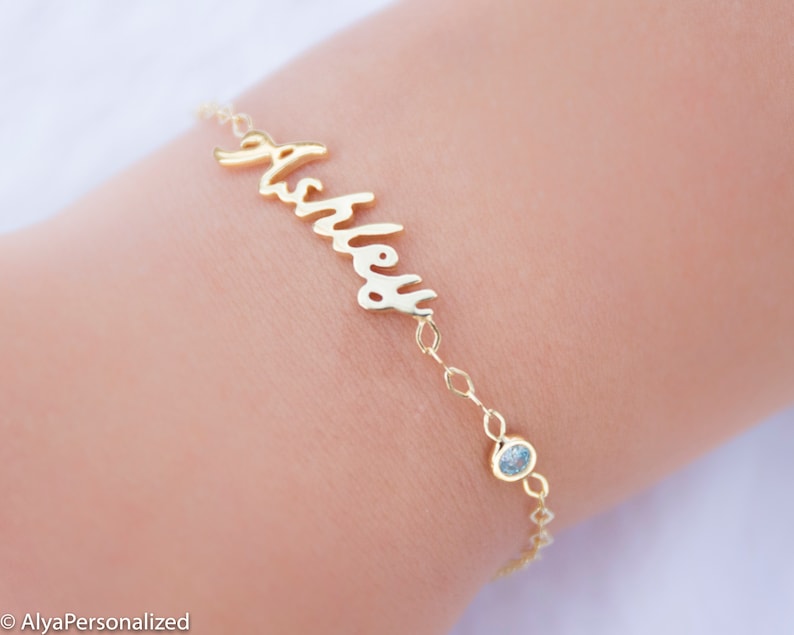 May include: Gold name bracelet with a light blue gemstone charm. The bracelet says "Ashley" in cursive.