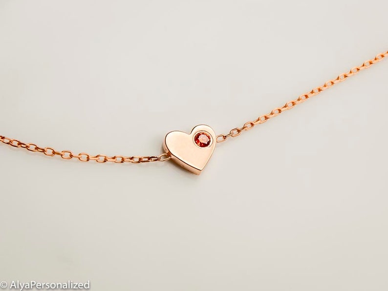 May include: A rose gold necklace with a heart pendant featuring a small red gemstone in the center.