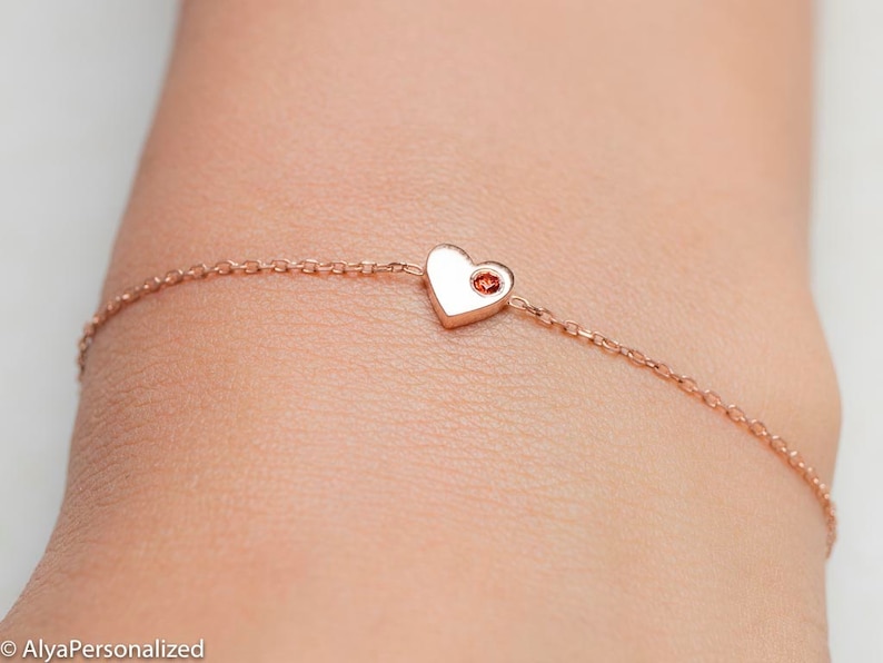 May include: A delicate rose gold chain bracelet with a small heart charm featuring a red gemstone in the center.