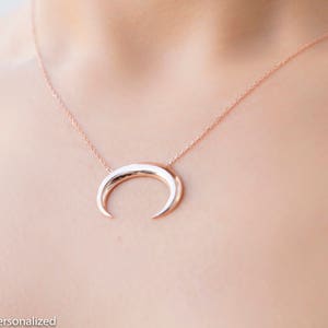 Può includere: Collana a mezzaluna in oro rosa. Il ciondolo ha una forma curva e lucida, sospesa da una delicata catena. La collana è mostrata su uno sfondo color carne.