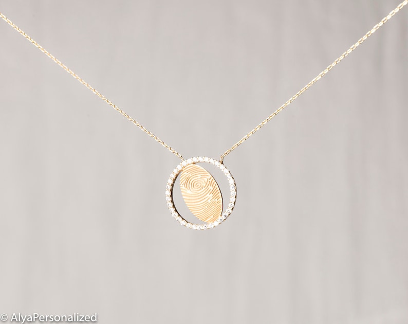 May include: A gold necklace with a delicate chain and a pendant featuring a fingerprint design surrounded by a circle of small diamonds.