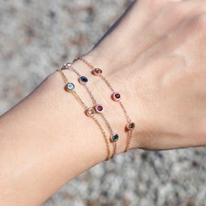 Puede incluir: Tres delicados brazaletes de cadena de oro con peque&ntilde;as piedras preciosas de colores. Cada brazalete presenta una piedra preciosa de color diferente, incluyendo azul, rojo y verde.