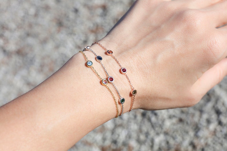 Puede incluir: Tres delicados brazaletes de cadena de oro con peque&ntilde;as piedras preciosas de colores. Cada brazalete presenta una piedra preciosa de color diferente, incluyendo azul, rojo y verde.