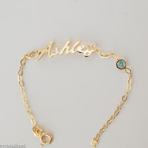 May include: Gold chain bracelet with a cursive nameplate that reads "Ashley" and a light blue gemstone.