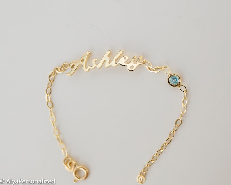 May include: Gold chain bracelet with a cursive nameplate that reads "Ashley" and a light blue gemstone.