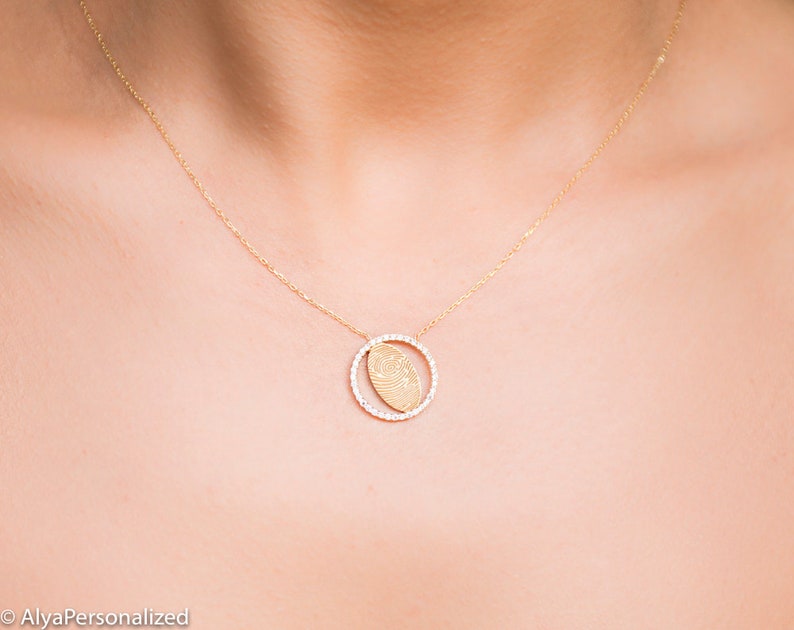 May include: A gold necklace with a delicate chain and a round pendant featuring a fingerprint design. The pendant is surrounded by a circle of small diamonds.