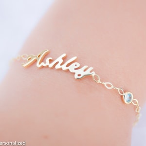 May include: Gold name bracelet with a cursive script name "Ashley" and a small clear gemstone charm.