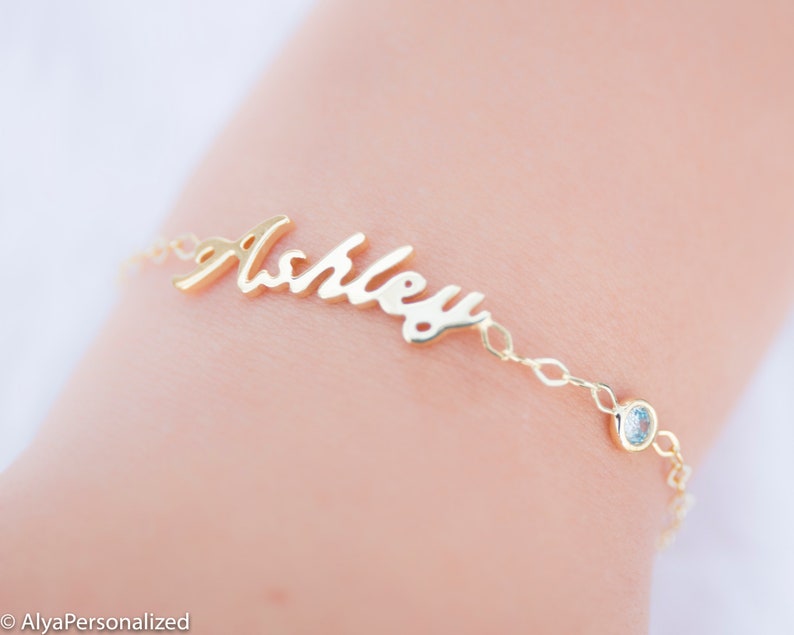 May include: Gold name bracelet with a cursive script name "Ashley" and a small clear gemstone charm.
