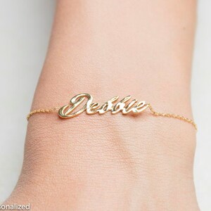 K&ouml;nnte beinhalten: Ein goldenes Kettenarmband mit dem Namen "Debbie" in Kursivschrift.