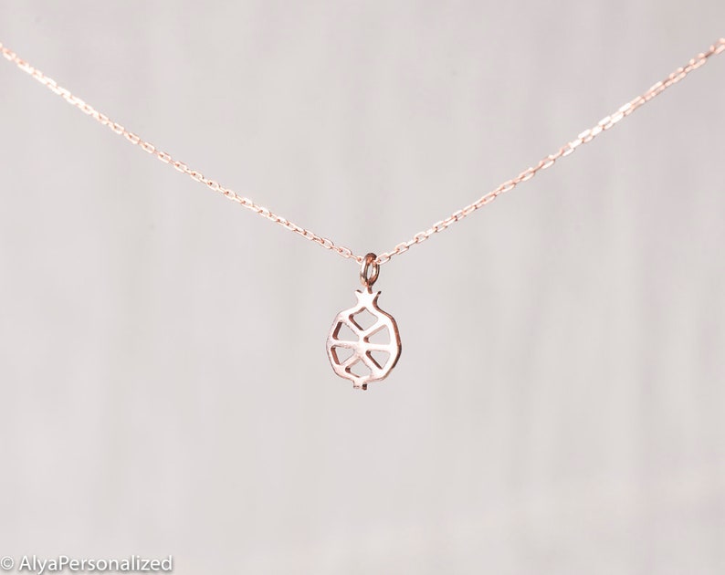 May include: A delicate rose gold necklace with a simple, geometric pomegranate pendant.