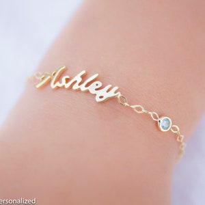May include: Gold name bracelet with a cursive script style name "Ashley" and a small blue gemstone accent.