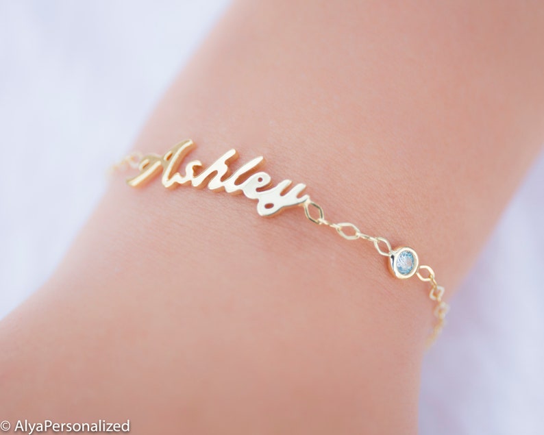 May include: Gold name bracelet with a cursive script style name "Ashley" and a small blue gemstone accent.