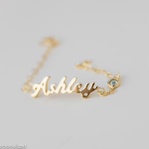 May include: Gold chain bracelet with the name "Ashley" in cursive script and a small blue gemstone.