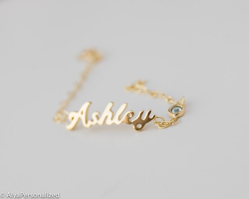 May include: Gold chain bracelet with the name "Ashley" in cursive script and a small blue gemstone.