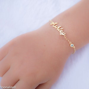 May include: Gold name bracelet with a blue gemstone charm. The bracelet is personalized with the name "Ashley".