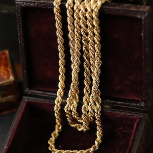 May include: A gold-colored rope chain necklace displayed in an open, antique jewelry box with a burgundy velvet interior. The chain is intricately woven, and the box is made of dark wood.