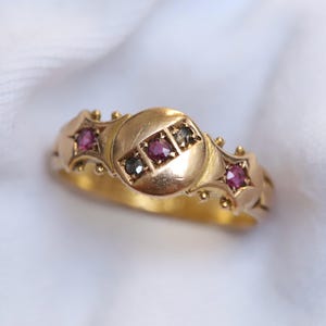 May include: A gold ring featuring a central circular plate set with a small red gemstone flanked by two dark stones. The band includes two additional star-shaped settings, each containing a single red gemstone, accented with small gold beads.